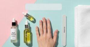 “A flatlay of natural nail-care items including cuticle oil, jojoba oil, a nail serum dropper, and a woman’s hand with short, healthy nails on a soft towel, styled on a pastel background.”